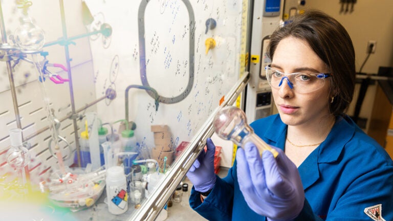 Student in blue lab coat holding and examining vial. In front of a scientific display with writing on the glass in front of her.