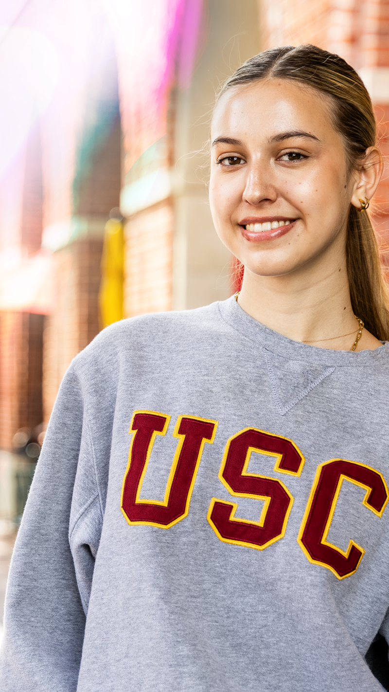 USC Dornsife female student looking at camera smiling. Wearing a grey USC sweater