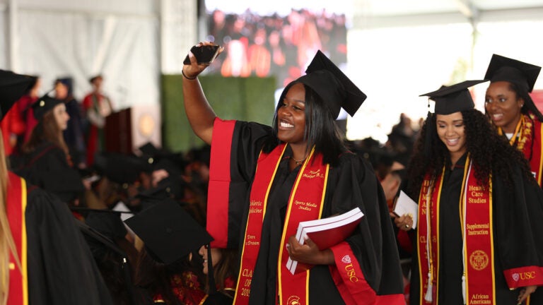 A woman at commencement wearing a cap and gown smiles and raises her hand.