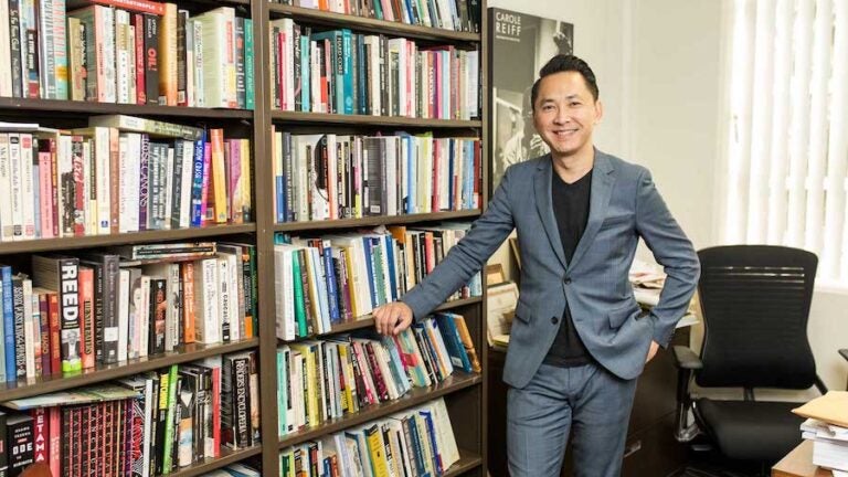 Viet Thanh Nguyen smiling to the camera while leaning on a bookshelf in an office