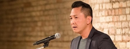 Professor Viet Nguyen speaking into a microphone in front of a brick wall