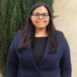 an image of Nelly Chavez with black-rimmed glasses and long black hair, in a navy blazer and smiling with teeth 