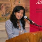 an image of Karina Casillas in medium-length black wavy hair, talking into a mic, with a light blue blazer and pearl necklace