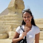 an image of Kendall Williams with the Cairo pyramids as the backdrop. She has long hair, is wearing a white short-sleeve shirt and is smiling with teeth