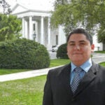 an image of David Hernandez in a black suit with a checkered black and white tie and light blue shirt. He has short hair and is smiling with no teeth