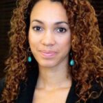 an image of Gina Clayton with medium length curly auburn hair. she has blue earrings and is wearing a black shirt. slight smiling with no teeth.