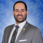 headshot of Oscar Rene Gutierrez in a gray suit and dark gray patterned tie. he has short hair, facial hair, and is smiling with teeth.