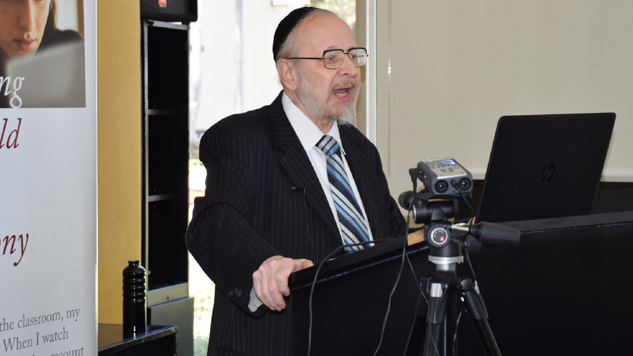 Harry Reicher speaks at his lecture from behind a podium. An audio recorder stands on a tripod by him.