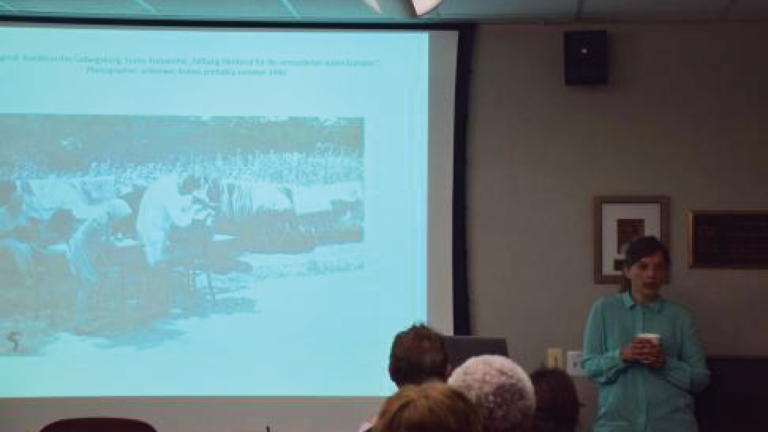 Julia Werner holds a cup of coffee while presenting her lecture in a History Department room. She is showing a historical photo on her projected presentation slides.