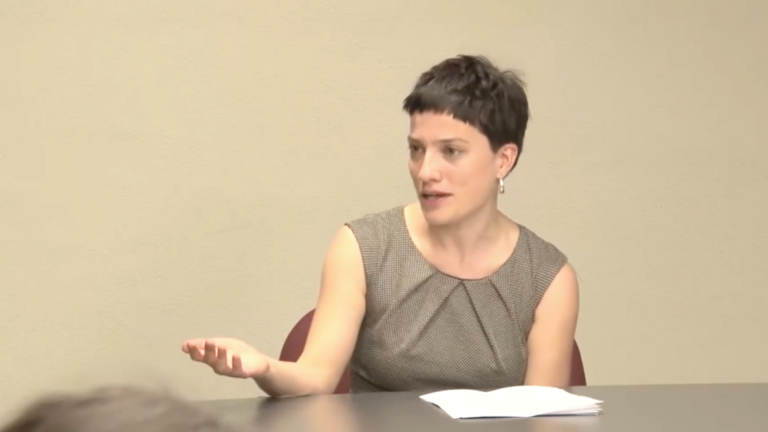 Screenshot from Katja Schatte's lecture video. She's sat at a table with a paper in front of her, gesturing and answering questions from the audience.