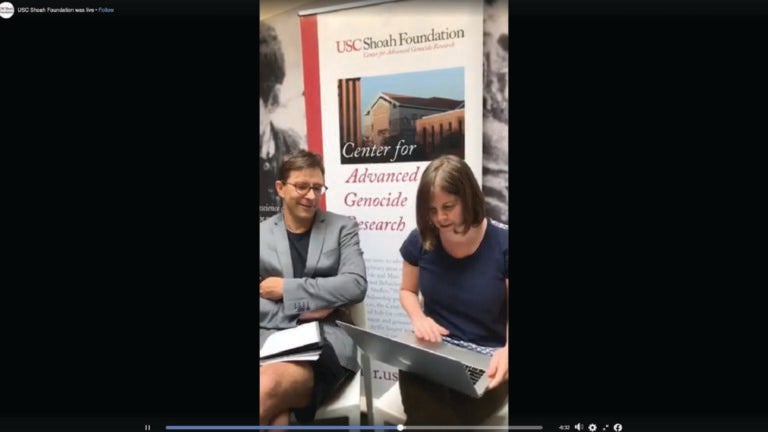 Screenshot of a Facebook live video. Alexander Korb, left, and an unknown staff member, right, sit in front of a center banner. Alexander has his arms and legs crossed. He smiles and looks down at the laptop the staff member is holding as they wait for questions from viewers.