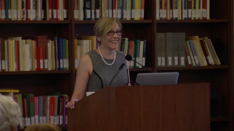 Screenshot of Diane Marie Amann presenting. She smiles, standing behind a podium and in front of a bookshelf filled with colorful books.