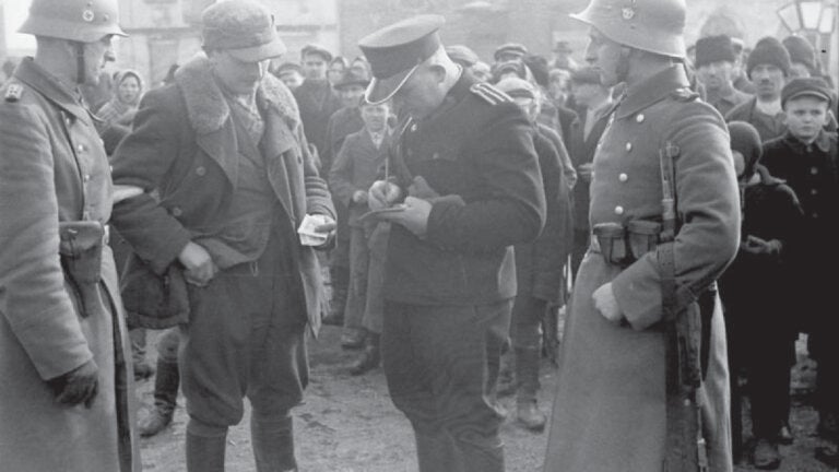 A black and white 1941 photograph depicting German law enforcement and Polish police officers checking a man’s documents in the Krakow district of Kazimierz in Poland. Text at the bottom reads Bundesarchiv Bild 101I-030-0781-07 Foto: Iffland 1941 Anfang.