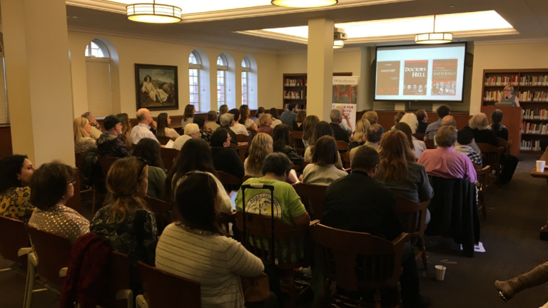A wide-shot of Diane Marie Amann lecturing in Doheny Library. A crowd of attendees sit in wooden chairs. Diane stands behind a podium.