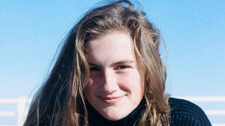 Cropped headshot of Virginia Bullington. Blue sky, a white fence, and the sea can be seen behind her.