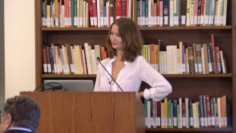 Bieke Van Camp stands behind a podium in Doheny Library with her arm on her hip. A bookcase full of books frames her in the background.