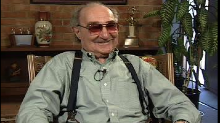 Screenshot from a Visual History Archive interview. An older white man smiles. He wears red tinted glasses, a grey-green button-up and suspenders.