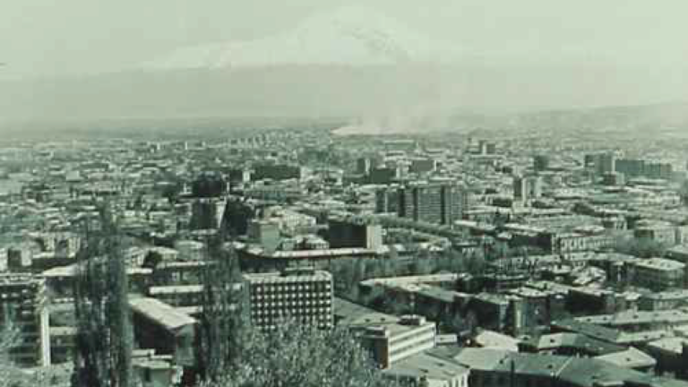 A 1960s photo of Mount Ararat in Armenia. In the foggy distance is the mountain, while in the foreground is the wide landscape of the city of Yerevan. Smoke or steam rises in the middle distance from a building. The photo is faded green with age.
