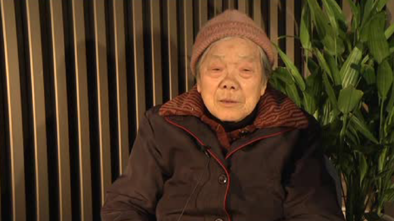 Screenshot from a Visual History Archive interview. An older Chinese woman speaks. She wears a coral pink knitted cap and a dark brown coat lined with red.