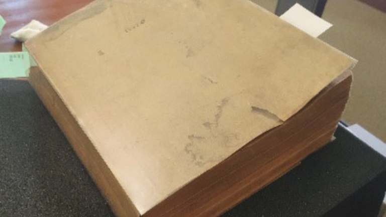 Picture of a thick, beige book with yellowed pages. A white piece of paper sticks out from the pages.