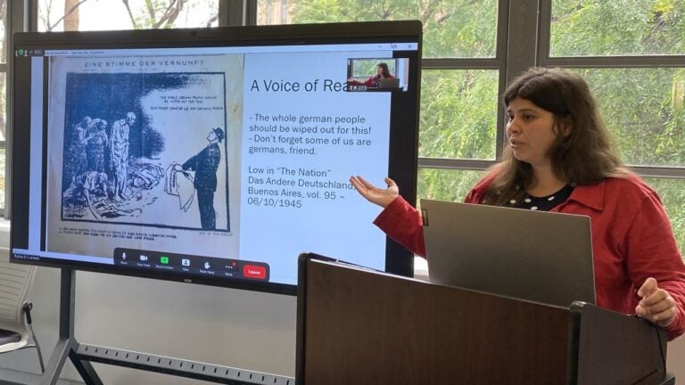 Raíssa stands at a podium and gestures to her presentation slide. It depicts an excerpt from a 1945 Buenos Aires newspaper, The Nation Das Anderes Deutschland.