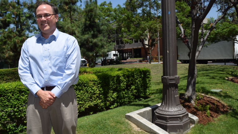 Adam R. Seipp poses for a picture on the USC campus. He smiles with his hands clasped at his waist, surrounded by greenery.