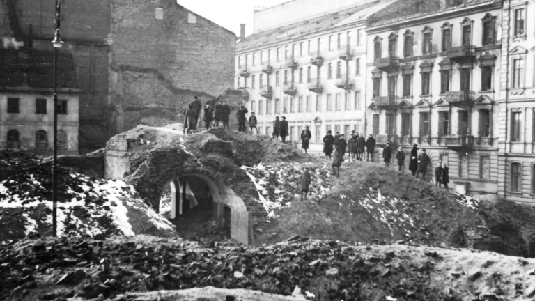 Historical black and white photo of the fuins of townhouses in Warsaw during WWII. A row of residents stand atop a mound of rubble, surveying the damage.