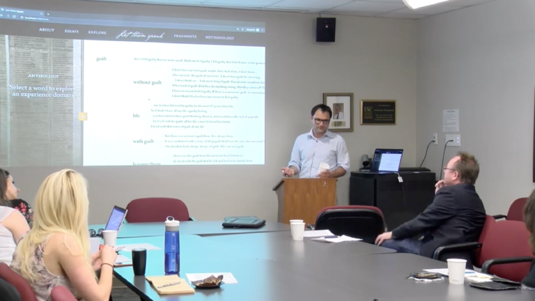A screenshot from Gabor Toth's lecture video. He gestures with a cup and pen in his hand in a History Department conference room.