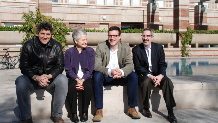 The Holocaust Geographies Collaborative smile against the bright sun, perched on the edge of Leavey Library’s fountain. They all look in different directions, and all have their hands clasped together in their laps. From left to right: Alberto Giordano, Anne Kelly Knowles, Tim Cole, and Paul Jaskot.