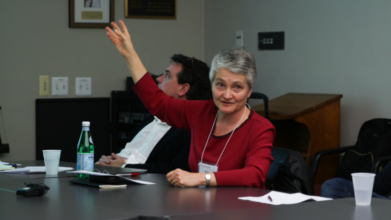 Anne Kelly Knowles raises a hand gesturing towards the projector slides out of frame. She is mid-speech, turned towards the other team members.