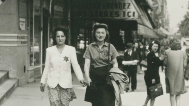 Young women wearing a Jewish badge stroll down a street in Paris during its Nazi-occupation. A woman in the background turns back to stare at them.