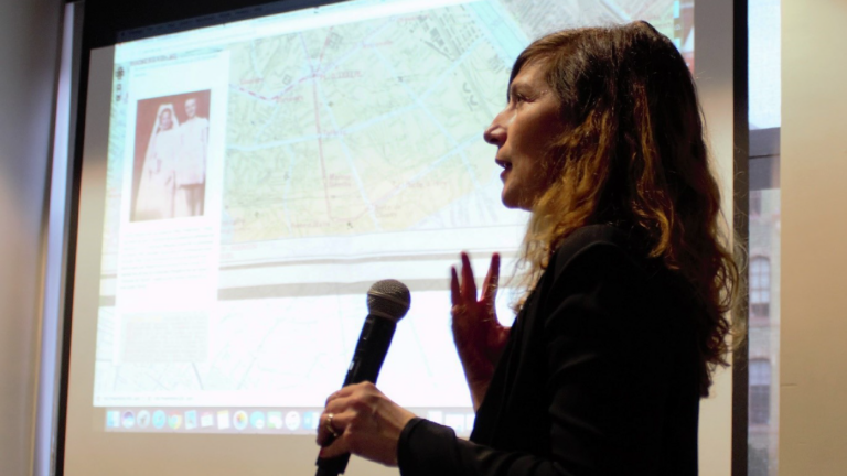 Mélanie Péron speaks at her lecture. She holds a mic in one hand and gestures with the other. A map and wedding photo can be seen on the slide projected behind her.