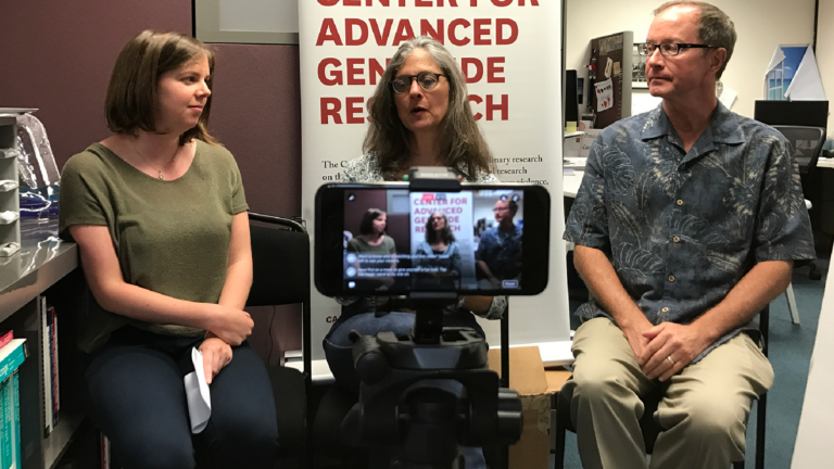 A Shoah Foundation staff member and fellows Nancy Sinkoff and Daniel Conway sit in front of a center banner. In the immediate foreground is a camera perched on a tripod, streaming the three to Facebook Live.