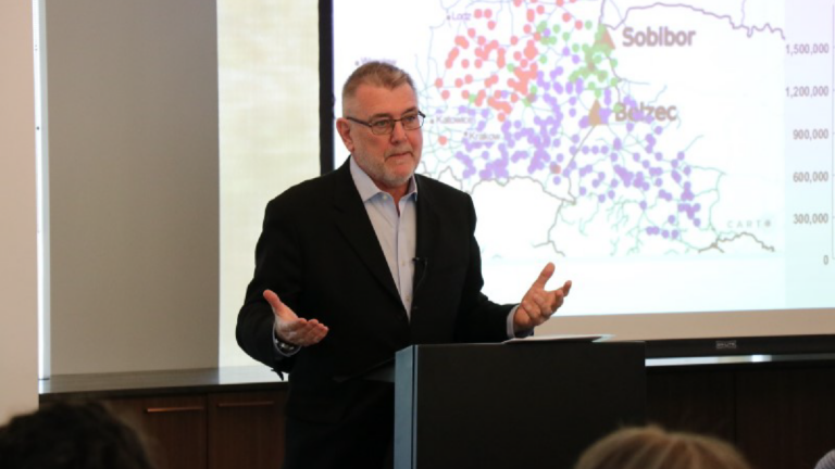 Photo of Peter Hayes lecturing. He has both hands out, palms up, gesturing. A map of Poland is projected behind him.