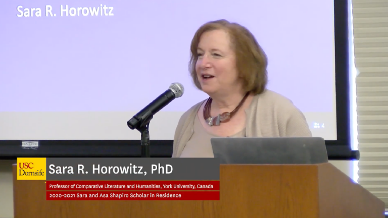 Screenshot of Sara Horowitz's lecture. She stands behind a podium and microphone. A digital banner displays her name, department, and fellowship.