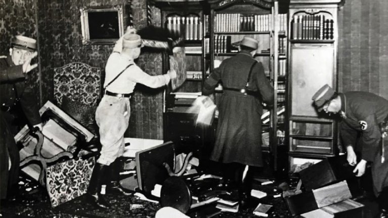 Nazi officials ransack a Jewish home, pulling books from a bookcase and throwing them on the floor. Overturned furniture and books litter the ground.