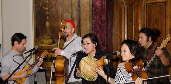 Five musicians performing in a band. There are three men with string instruments and two women. One woman is using an Afoxé and the other woman on a violin.