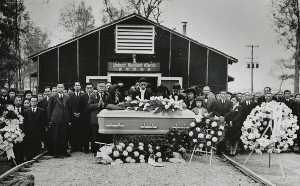 Various people wearing black gathered around a casket outside of a Buddhist church.
