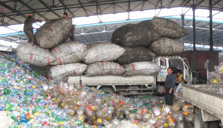 A large pile of plastic bottles and containers. Workers in the back are unloading giant sacks from a truck.