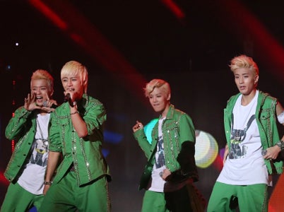 A male K-pop group performing on a stage. They are all wearing matching green jackets and pants.