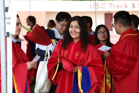 A PhD student stands in cap, hood, and gown at commencement. She smiles into the camera as other students move around her in the background.