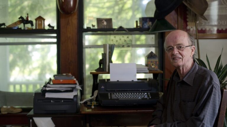 Author James Tate sits at the right of this image. In his writing studio, a typewriter, books, and small trinkets frame the background.