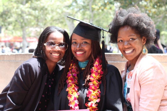 A family of three women surround a new alumni student in cap and gown.