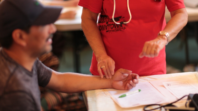 A person with a stethoscope takes the pulse on the wrist of another person seated at a table