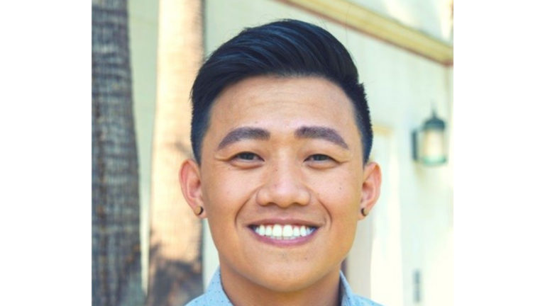 Headshot of Thai Le, a Vietnamese man with slick back hair, wearing a light blue dotted button down shirt and earrings, smiling brightly
