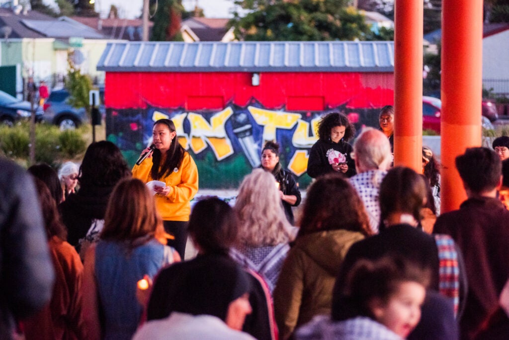 Community members gather for an APEN youth-led vigil holding candles. A young female with a yellow sweater stands at the front of the crowd holding a microphone to speak.