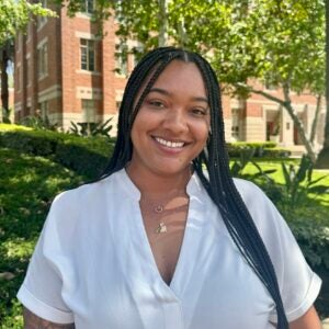 A headshot of a Black woman with their hair in long, thin braids, wearing a white blouse posing with a smile in front of a building and trees on the USC campus.