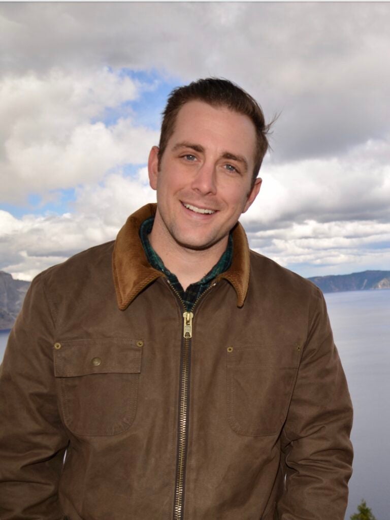 Portrait of Jameson Karns in a brown jacket in front of body of water with hills in background and cloudy sky.