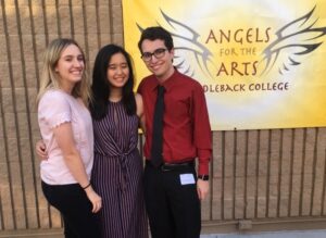 Three students posting in front of a sign saying "Angels for the arts"