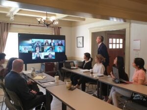 A man stands and other people sit around a table while conducting a video call with several people on a monitor